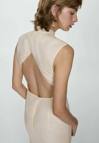 Beiges ärmelloses Kleid mit offenem Rückendesign, hoher Halslinie und einem Reißverschlussdetail im unteren Rückenbereich. Glatte Stofftextur.