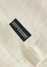 Étiquette en tissu noir avec texte blanc "SUPERDRY" cousue sur tissu texturé blanc cassé avec couture visible.