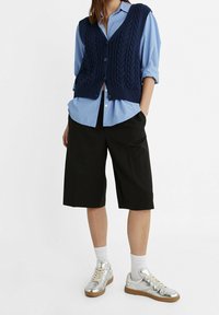 Mörkblått stickat väst med kabeldesign över en blårandig skjorta, matchat med svarta knälånga shorts och silverfärgade sneakers med brun sula.