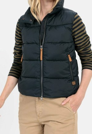 Kvinde iført sort dunvest med lynlås og lysebrune lynlåstræk over en sort og olivengrønstribet langærmet skjorte og beige bukser.