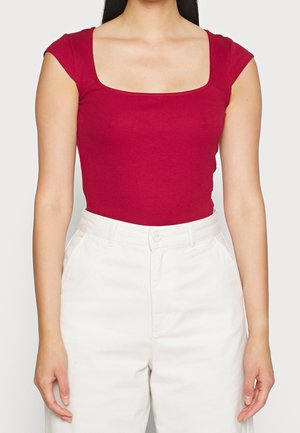 Frau trägt ein eng anliegendes rotes Kurzarmoberteil mit quadratischem Ausschnitt und weiße Hose mit hoher Taille, steht vor einem schlichten Hintergrund.