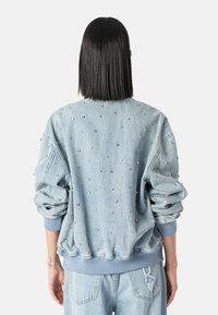 Osoba s rovnými černými vlasy, oblečená v světle modré džínové bundě a džínách, bunda zdobená malými stříbrnými nýty, stojící zády.