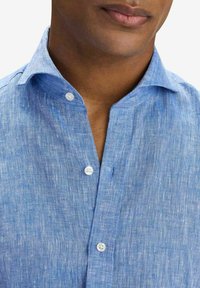 Ljusblå linneskjorta med krage, som har en knäppning och två vita knappar. Tyget har ett strukturerat, vävt utseende.
