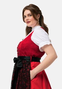 Robe rouge et noire avec des motifs complexes. Corsage texturé, chemisier blanc en dessous, ruban en satin à la taille, et poches dans la jupe. Des pois sont également présents.