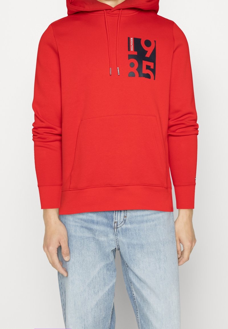 Sweat à capuche rouge avec poche avant et motif noir "1985" sur la poitrine, porté avec un jean bleu clair par une personne debout.