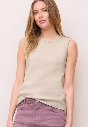 Jeune femme aux longs cheveux blonds portant un haut beige sans manches et un pantalon taille haute mauve, debout devant un fond uni.