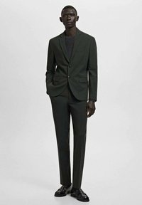 Mężczyzna w ciemnozielonym garniturze z dwuguzikową marynarką i dopasowanymi spodniami, w czarnych skórzanych mokasynach, stoi na jasnym tle.