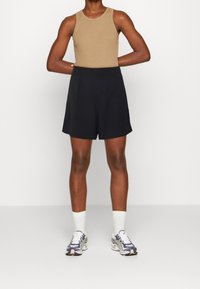 Beżowy top na ramiączkach bez rękawów i czarne szorty. Model nosi białe skarpetki i szaro-niebieskie sneakersy. Prosty design, wygodny krój, z luźną sylwetką.