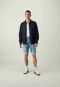 Mladý muž v černé košili na knoflíky přes bílém tílku, s krátkými světle modrými denimovými šortkami, bílými sockami a bílými teniskami stojí před jednobarevným pozadím.