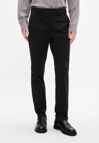 Pantalones negros de tela suave, con corte recto, trabillas para el cinturón y cierre de botón. Combinados con zapatos negros y una camisa gris.