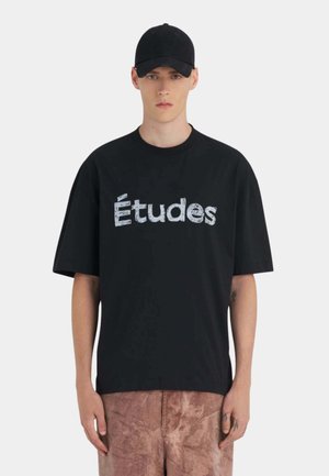 Jeune homme portant une casquette noire, un T-shirt noir surdimensionné avec le texte "Études" et un pantalon marron texturé, debout devant un fond clair uni.