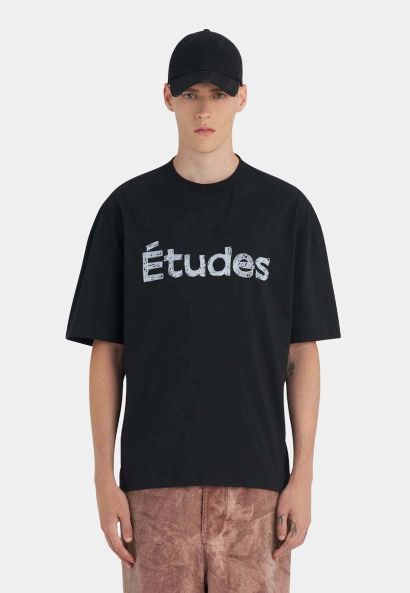 Jeune homme portant une casquette noire, un T-shirt noir surdimensionné avec le texte "Études" et un pantalon marron texturé, debout devant un fond clair uni.