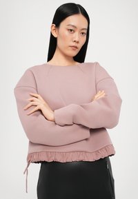 Un hanorac roz scurt, cu o margine volan, mâneci lungi și o textură netedă. Modelul poartă hanoracul cu brațele încrucișate.
