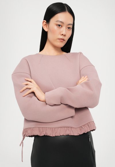 Rosa beschnittenes Sweatshirt mit volantrippeligem Saum, langen Ärmeln und glatter Textur. Model trägt das Sweatshirt mit verschränkten Armen.