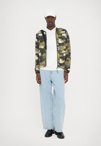 Camo zip jacket in green and beige, white hooded sweatshirt, light blue wide-leg jeans, and black shoes, standing against a white background.