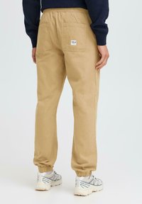 Beige pamut joggers, rugalmas derekkal, bordázott szegélyekkel és két hátsó zsebbel. A jobb hátsó zsebben címke található.