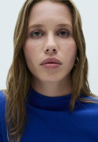 Pogled od blizu na osobu s dugom, valovitom smešnom kosom koja nosi plavi top s visokim ovratnikom. Osobine uključuju jasnu kožu i neutralnu boju usana na svetloj pozadini.