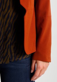 Blazer color ruggine con trama liscia sopra una camicia a strisce blu navy e verde oliva. Jeans in denim scuro visibili in basso. Nessun hardware distintivo.