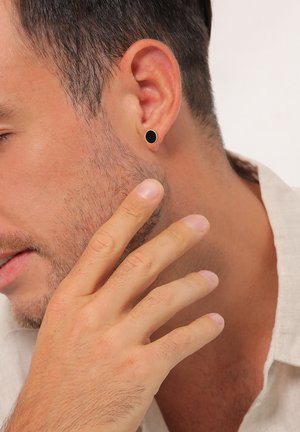 Schwarze Stud-Ohrringe, aus Metall, runde Form, minimalistisches Design. Getragen am linken Ohr, vor einem hellen Hautton und einem stoppeligen Kiefer.