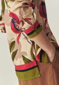 Mouwdetail van een bloes met bloemenprint met rode, groene, en zwarte accenten, knoopmanchet, en hand in de zijzak van een bruine broek.