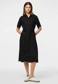 Juoda suknelė, su sagomis priekyje, trumpomis rankovėmis ir rišamu juosmens dirželiu. Lygus audinys, iki kelių ilgio su klasikinio tipo apykakle.