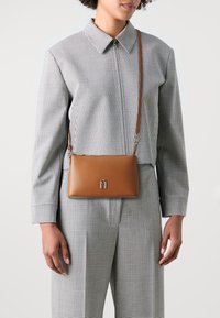 Borsa a tracolla in pelle marrone con un'accentuazione di logo argentato, caratterizzata da una superficie liscia e una forma rettangolare, indossata sopra una camicia a quadri.