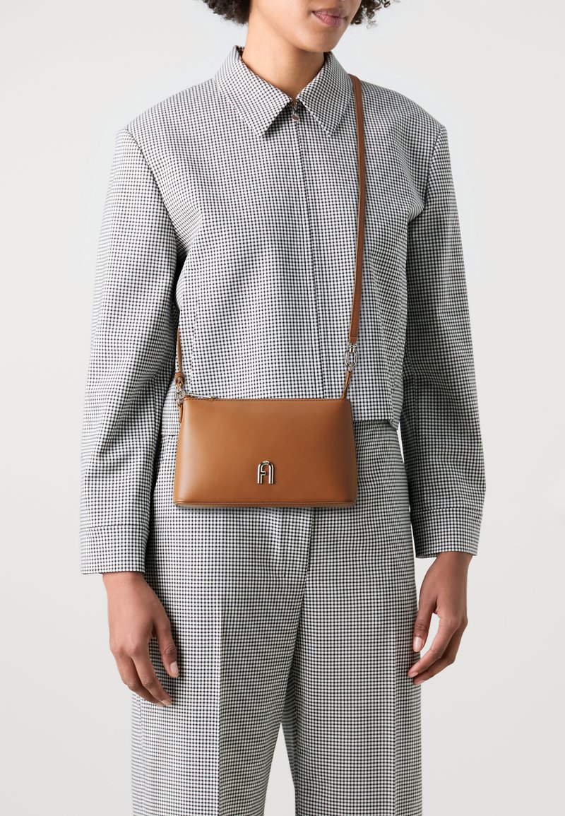 Borsa a tracolla in pelle marrone con un'accentuazione di logo argentato, caratterizzata da una superficie liscia e una forma rettangolare, indossata sopra una camicia a quadri.