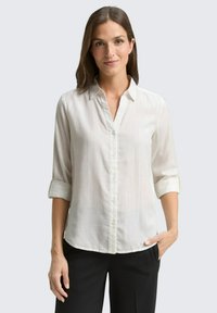 Chemise boutonnée beige clair avec un col, manches retroussées et une texture verticale subtile, assortie à un pantalon noir.