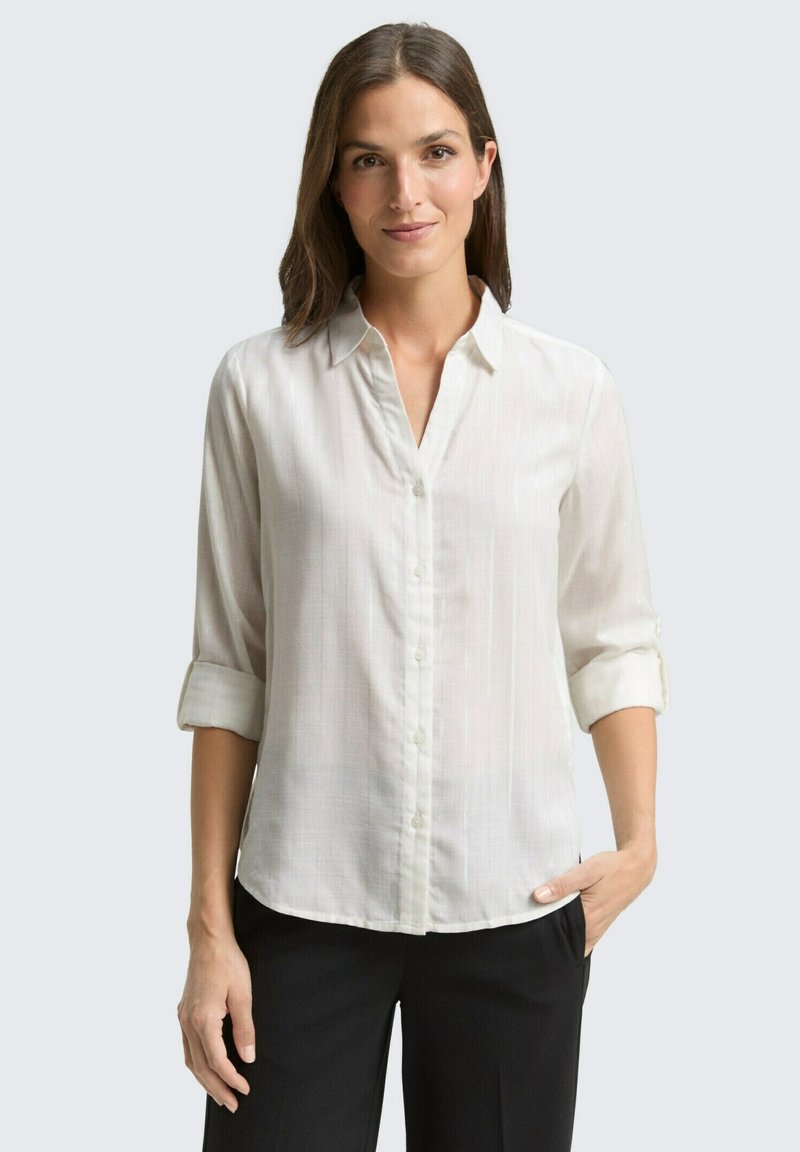 Chemise boutonnée beige clair avec un col, manches retroussées et une texture verticale subtile, assortie à un pantalon noir.