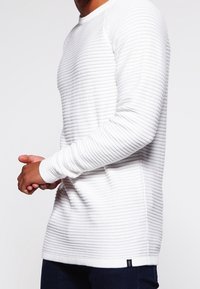 Maglione bianco a maniche lunghe con texture e righe orizzontali. Presenta un collo rotondo e polsini aderenti. Piccolo logo in basso a sinistra.