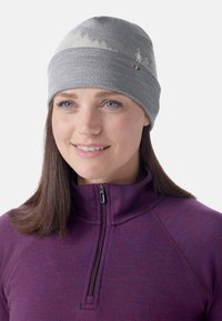 Grå strikket hue med bjergmønster og kontrastfarvede accenter. Bæres med en lilla zip-neck top i struktureret stof.