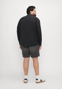 Schwarzes Langarmhemd, graue Denim-Shorts, weiße Sportschuhe mit schwarzen Akzenten. Von hinten gesehen, zeigt den Kragen und die Knöpfe des Hemdes.