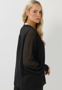 Blusa nera trasparente con motivo a pois testurizzati, maniche lunghe e polsini arricciati. Tessuto leggero con una vestibilità rilassata.