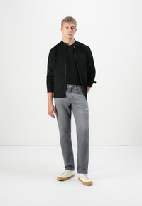 Joven de pie, vistiendo una chaqueta negra sobre una camisa negra, jeans grises con ligero desgaste y zapatillas blancas con suelas de goma.