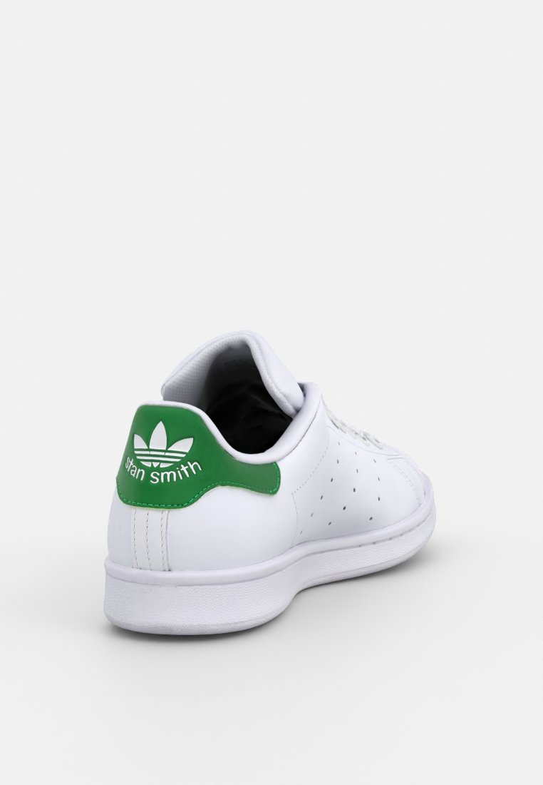Baskets blanches avec un talon vert, design perforé et semelle blanche plate. Présente une marque "Stan Smith" à l'arrière. Texture en cuir lisse.