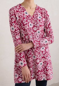 Czerwona tunika w kwiaty z dekoltem w serek, długimi rękawami i skomplikowanymi białymi oraz niebieskimi wzorami kwiatowymi. Miękka, teksturowana tkanina.