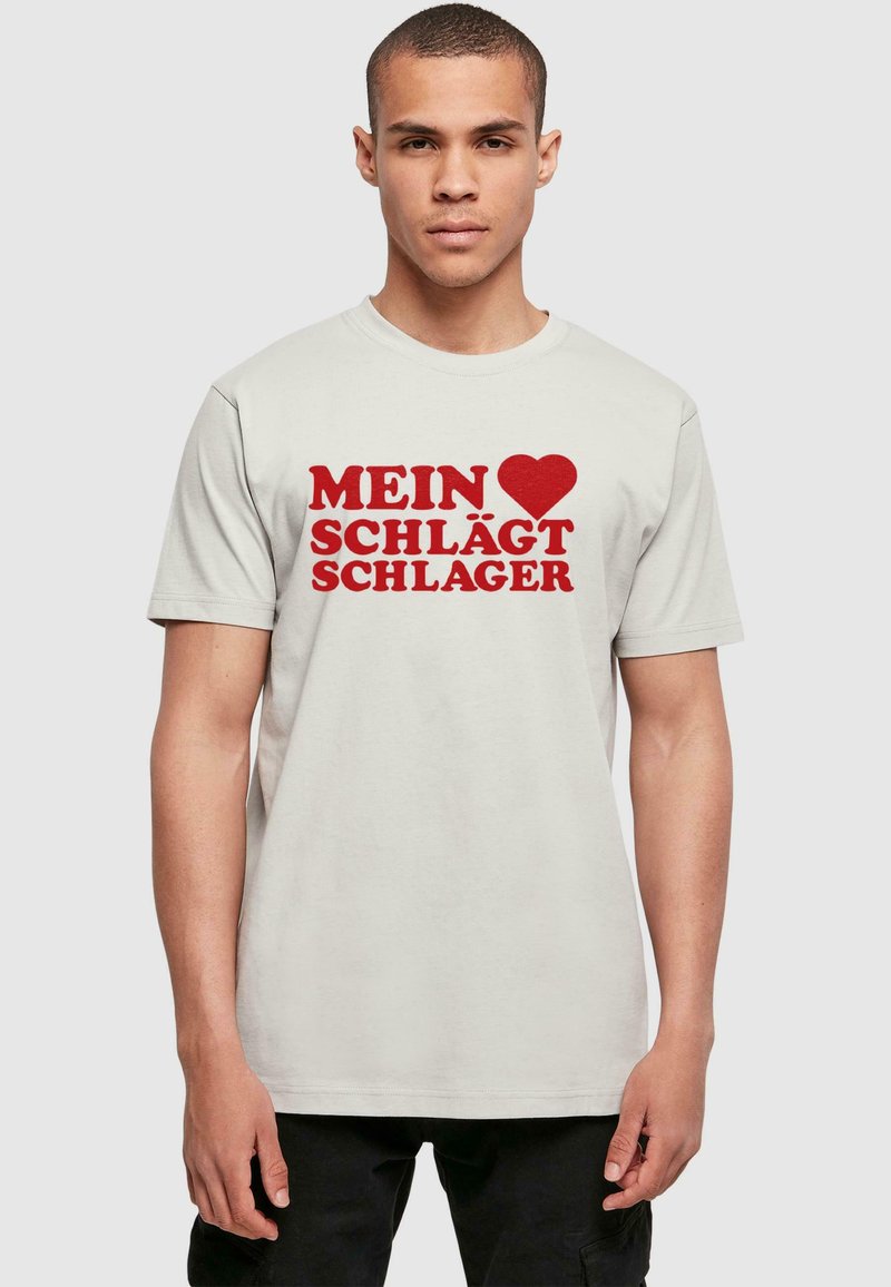 Lys beige t-shirt med korte ærmer, der har fed rød tekst, som står "MEIN ♥ SCHLÄGT SCHLAGER," rund halsudskæring og afslappet pasform.