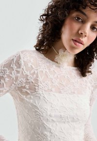 Weißes florales Spitzenoberteil mit einer figurbetonten Silhouette, das über kunstvolle Stickerei, transparente Textur und lange Ärmel verfügt. Akzentuiert mit einem blumigen Choker.