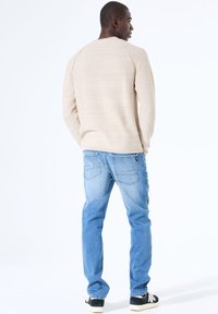 Beige textuurtrui met een ronde hals, gecombineerd met lichtblauwe jeans en zwarte sneakers, gezien vanuit de achterkant.