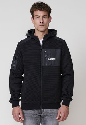Sudadera con cremallera negra fabricada en tejido texturizado, con un panel liso en el pecho, bolsillo con cremallera en el brazo y capucha con cordón.