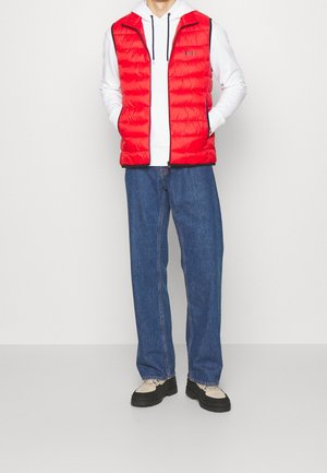 Veste matelassée rouge avec fermeture éclair, col montant et bordure contrastante foncée, portée sur un sweat à capuche blanc, associée à un jean en denim bleu clair et des baskets noires.