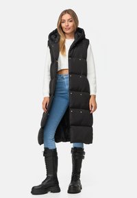 Svart stoppad lång väst med huva, med guldknappar och quiltad textur. Bärs över en vit kortärmad tröja och blå jeans.