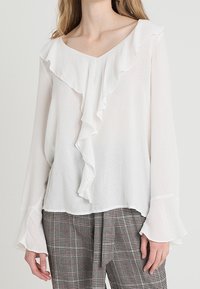 Blusa bianca con scollo a V arricciato e maniche lunghe a campana, realizzata in tessuto leggero; indossata con pantaloni a quadri con un motivo grigio e rosso.