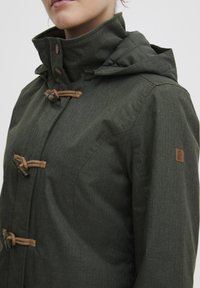 Dark green jacket with a textured fabric, featuring a hood, toggle closures with rope accents, and a small logo patch on the sleeve.