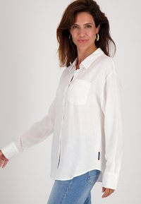 Vrouw met lang bruin haar, draagt gouden oorbellen, een losse witte blouse en lichtblauwe jeans, staat zijwaarts op een witte achtergrond.