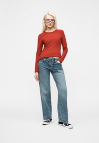 Νεαρή γυναίκα που φοράει μαύρα γυαλιά, κόκκινη μπλούζα με μακριά μανίκια, μπλε τζιν με κορδόνια και μαύρα αθλητικά παπούτσια, στέκεται μπροστά σε έναν απλό λευκό φόντο.