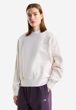 Jonge vrouw met steil bruin haar, draagt een wit sweatshirt en paarse joggingbroek, staat met één hand in de zak.