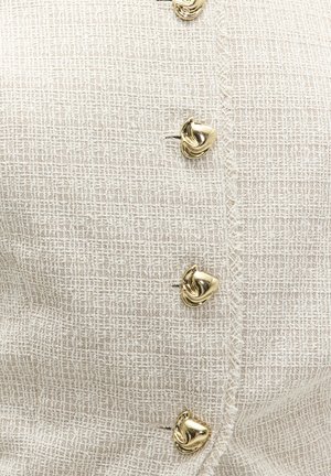 Tissu en crêpe tissé crème avec rangée verticale de quatre boutons en métal doré torsadés, bords texturés le long de la couture des boutons.