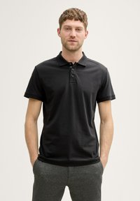 Sort polo t-shirt med korte ærmer, tre-knaps knapliste, glat tekstur, båret med grå slim-fit bukser. Har et klassisk kravedesign.