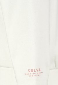 Witte sweatshirt met ribgebreide manchetten en onderzoom. Kenmerkt zich door de roze tekst "SBLVL CONTEMPORARY CLOTHING" aan de linkerkant. Zachte textuur, casual ontwerp.